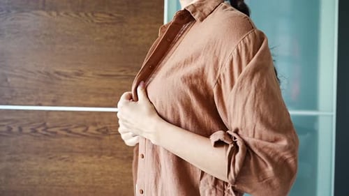 Cropped View of Young Woman Putting on Brown Shirt and Fastening Buttons in Front of Wardrobe