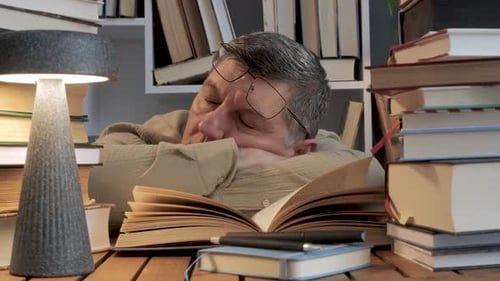 Exhausted Man Sleeping at Desk Surrounded By Books During Study