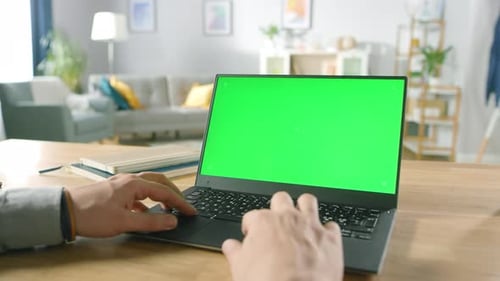Close-up of a Man Uses Laptop with Green Mock-up Screen While Sitting at the Desk in His Cozy Livin
