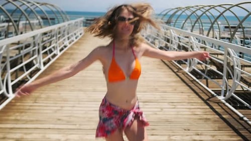 A woman in swimsuit on wooden pier dances to rhythm of music on sparkling turquoise sea water