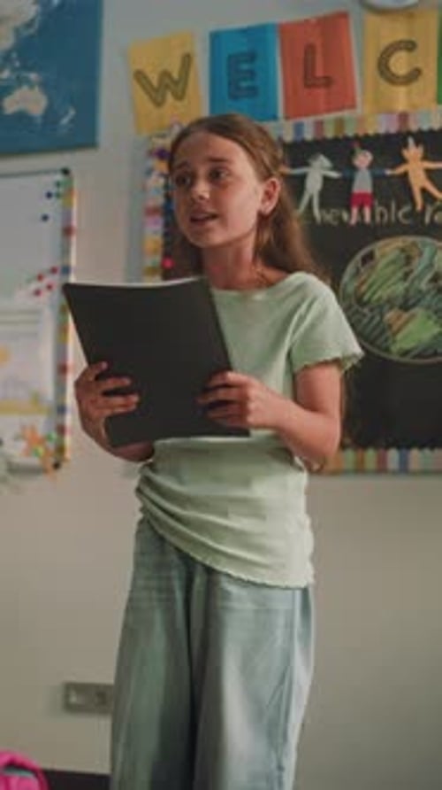 Primary School Girl with Notebook Showcasing Knowledge of Ecology in Front of Classmates and Teacher