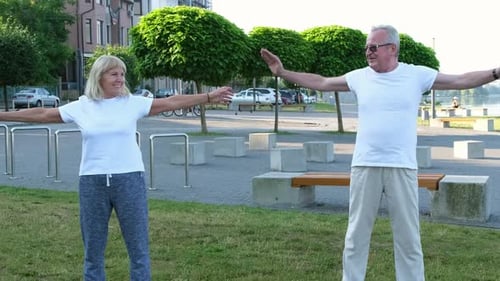 An Elderly Couple is Doing Sports Outdoors Stretching in the Park at Sunrise