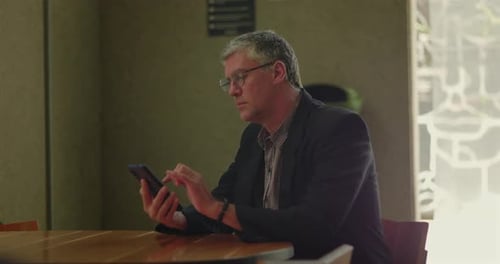 Mature man with gray hair and eyeglasses using smartphone while seated at wooden table in office