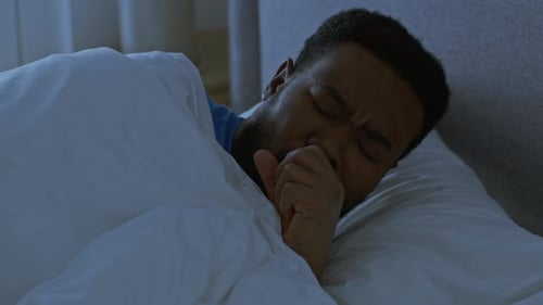 Junger schlafender Afroamerikaner wacht wegen eines Hustenanfalls auf und liegt spät in der Nacht im Bett