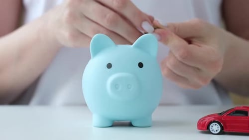 Woman Putting Coins in Blue Piggy Bank with Red Car Model on Foreground