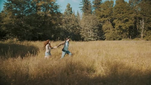 A young couple are holding hands while running through a tall grass field.