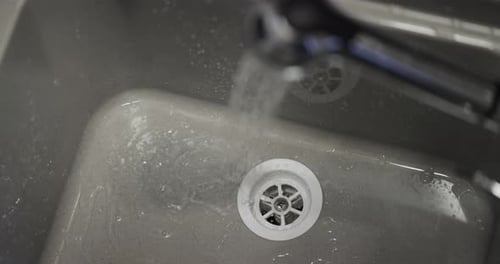 Close-up of water running from a faucet into a grey plastic sink with a white drain, 4K