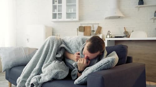 Young man trembling from fever. Sick man lying on couch at home and covering