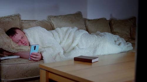 A man with a mobile phone in his hand lying on the sofa in front of the TV, covered with a blanket