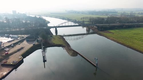 A bridge spans a calm river in a rural landscape, with fields, trees and a small riverside work area