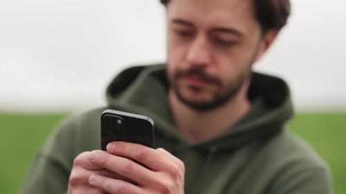 A Brunette Man Uses a Phone While Sitting in a Park A Bearded Man in a Green Jacket is Typing