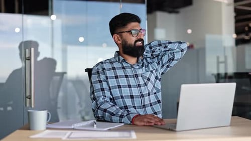 Tired Indian Businessman Stretching Neck at Desk in Modern Office Workplace Wellness Concept