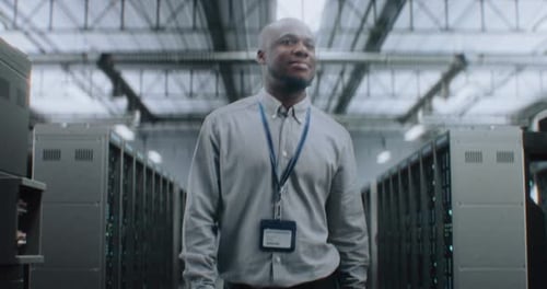 Big Data Center with Server Racks Professional Specialist Walking In Warehouse Looking at Camera