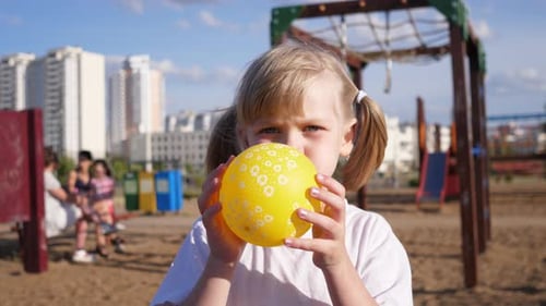 Little Girl Child Portrait Inflates a Yellow Balloon on the Playground in Summer