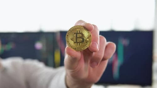 Hand Holds Gold Bitcoin in Front of Computers