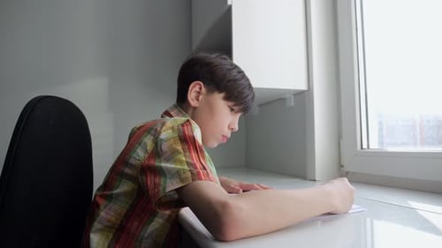 Teen Schoolboy Writes Homework in Notebook at Table in Room