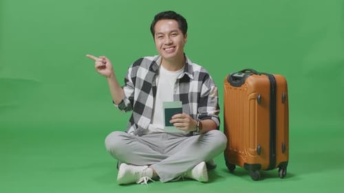 Asian Male Traveler With Luggage And Passport And Pointing To Side While Sitting In The Green Screen