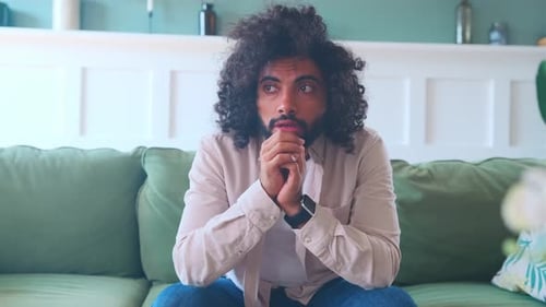 Young Perplexed Arabian Man Thinks and Holds Hands Close to Chin Sits on Sofa