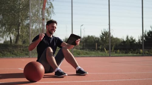 Man Sits on Basketball Court and Looks at Tablet and Rejoices at Winning Bet