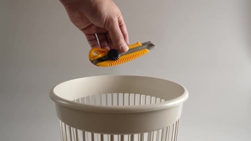 Hand Drops Yellow Box Cutter into Trash Can