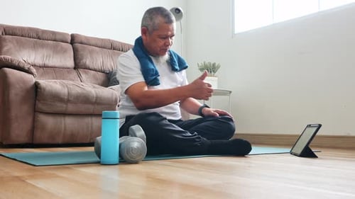 Elderly man follows exercise movements demonstrated by online trainer on yoga mat