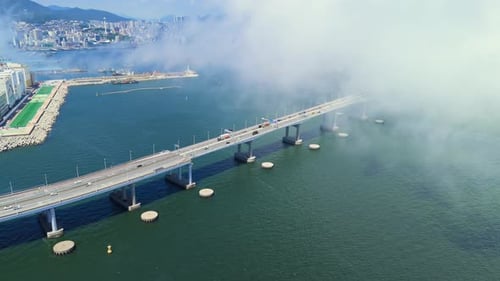 Aerial of a Concrete Bridge Over Green Ocean Water Shrouded in Fog with a City Skyline and Mountains