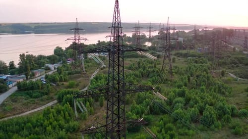 Structure of high-voltage lines outside the city at sunset. Trees and river. Camera motion back.