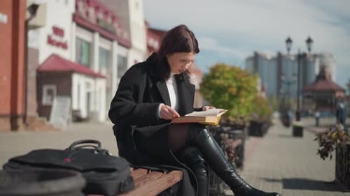 Young Lady Seated on Bench Reading Book Outdoors with City Background