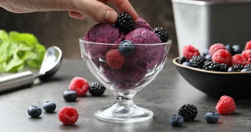 Woman putting berry and mint onto delicious sorbet at grey textured table against dark background