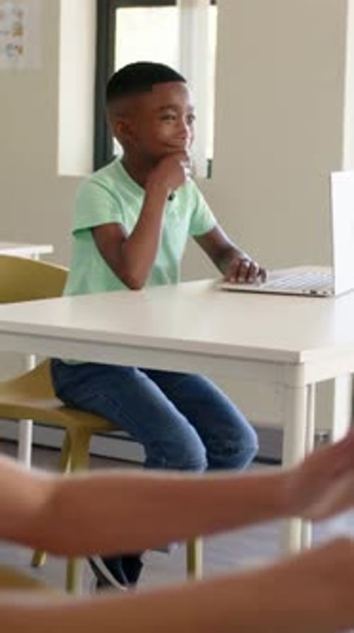 Vertical video: Camera shifting focus, blurring boy following teacher at classroom desk with laptop