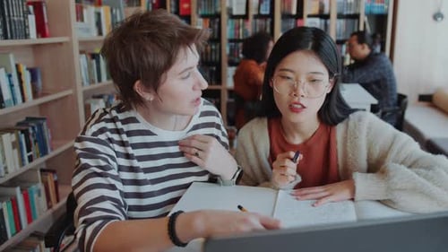 Two Diverse Female Students Discussing Information on Laptop in Library