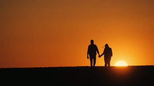 Händchen haltend geht ein junges Paar zusammen über ein Feld und bewegt sich der untergehenden Sonne entgegen