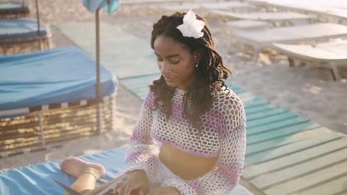 Afroamerikanische Frau arbeitet an einem Laptop, während sie bei Sonnenuntergang am Strand sitzt, umgeben vom Strand