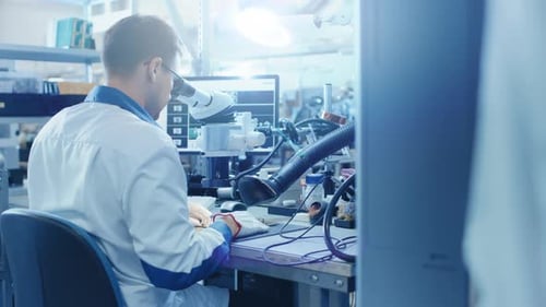 Electronics Factory Worker in White Work Coat Inspects a Printed Circuit Board Through a Digital Mi
