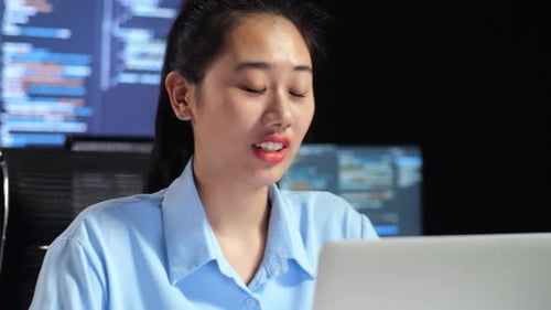 Asiatische Programmiererin bei einem Meeting an einem Laptop, während sie Code auf mehreren Monitoren schreibt