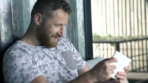 Young Man Playing Game on Tablet and Relaxing on Sofa in Cafe 30s