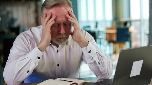 Troubled senior man sits at desk holding by the head. Tired overworking businessman in office.