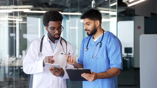 Two doctors standing in modern hospital discussing patient information while using digital tablet