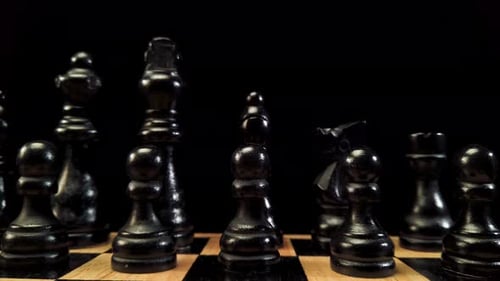 Truck shot from right to left of black chess pieces standing on a chessboard on a black background.