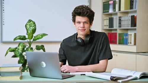 Portrait of College Student Guy Sitting at Desk with Laptop Inside an Educational Library