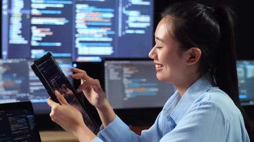 Side View Of Asian Female Programmer Looking At Database On Tablet While Writing Code By A Laptop