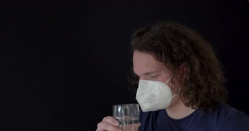 Young white curly man trying to drink water wearing mask with black background