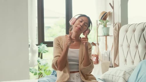 Asian girl wear white cream pajamas wear pink headphone on fluffy comfort bed happy dancing listen.