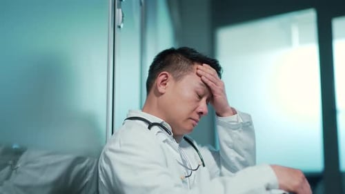 stressful exhausted Asian doctor sit in the hallway indoors a hospital. Tired pensive thoughtful
