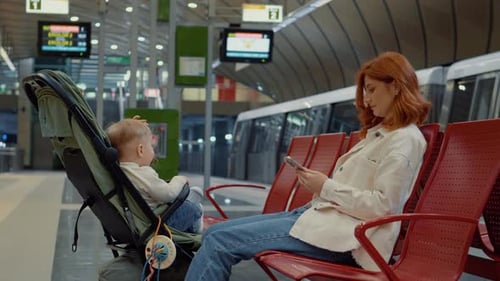 Young mother waits for subway with baby while scrolling on her phone