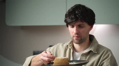Man Eats a Cake and Drinks Tea for Breakfast Sitting at a Table in the Kitchen