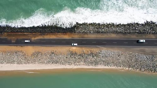 Aerial drone shot, top-down view of a road flanked by ocean waters on both sides, with waves washing