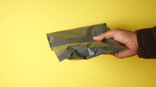 Hand with a Crumpled Black Package on a Yellow Background