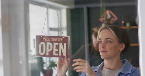 Smiling caucasian female hairdresser turning shop sign to open on door of hair salon, in slow motion