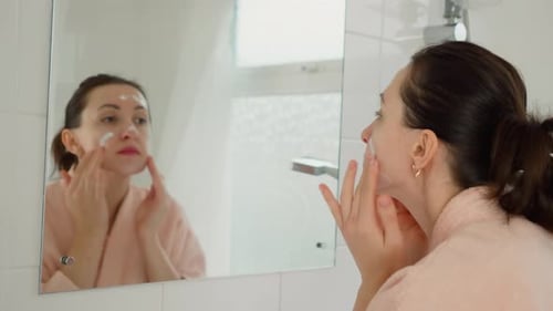 Woman Applying Facial Cream in Bathroom Mirror
